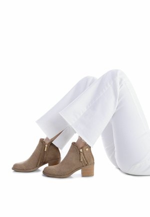 Botines marrones perforados con tacón bajo de bloque y cremalleras laterales, combinados con pantalones acampanados blancos. Superficie limpia y texturizada.