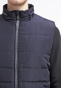 Granatowa pikowana kamizelka z wysokim kołnierzem, zamkiem błyskawicznym z przodu i przeszywanymi poziomymi panelami; w połączeniu z teksturowaną czarną koszulą z długim rękawem.