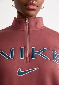 Sweatshirt cor de bordô com gola alta, fecho de meia-zíper e logotipo "NIKE" bordado em azul-marinho e branco. Textura macia, corte casual.