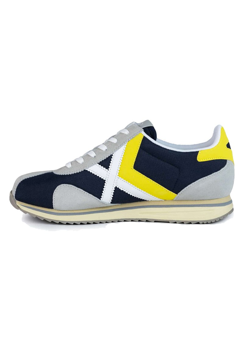 Zapatillas azul marino y gris con acentos blancos y detalles en amarillo. Presentan un tejido texturizado y una suela de goma con un patrón en zigzag.