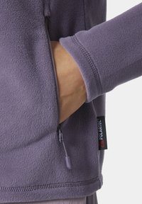 Fioletowa polarowa kurtka z zamykaną na zamek boczną kieszenią i metką Polartec. Charakteryzuje się miękką teksturą i dopasowanymi mankietami, zaprojektowana z myślą o ciepłe i komforcie.
