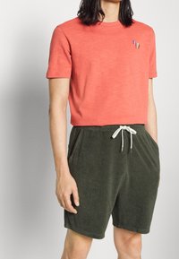 Chemise corail à manches courtes avec logo brodé multicolore sur la poitrine, associée à un short en éponge vert foncé avec cordon blanc.