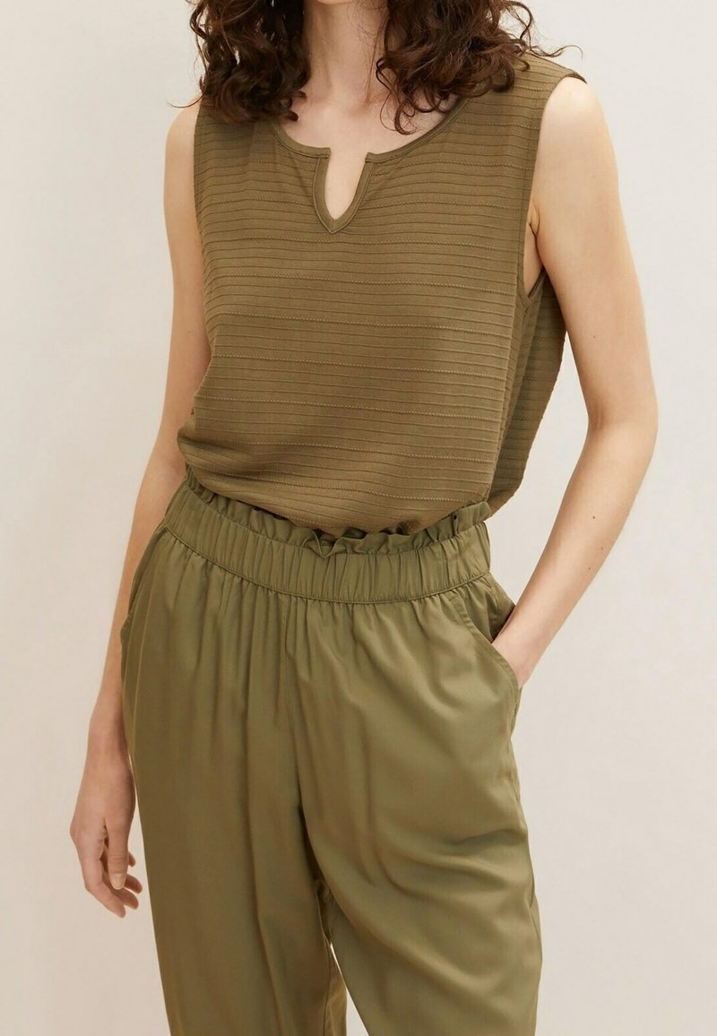 Femme portant un haut sans manches en côtes vert olive avec une encolure en V et un pantalon assorti à taille élastique, une main dans la poche.
