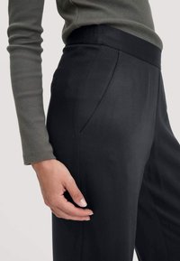 Zwarte broek met een gladde textuur, voorzien van zijzakken en een aansluitende tailleband. Een hand rust zachtjes op de stof.
