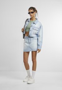 Gaiši zila saīsināta džinsu jaka ar priekšējām kabatām, apvienota ar zaļu saīsinātu topu un džinsu mini svārkiem. Balti sporta apavi un saulesbrilles noslēdz izskatu.