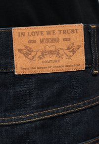 Parche de cuero en jeans de mezclilla oscura con la inscripción "EN EL AMOR CONFIAMOS MOSCHINO COUTURE" y dos pájaros sosteniendo una pancarta con un corazón.