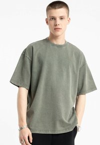 Przestronny oliwkowy t-shirt z bawełny z krótkimi rękawami, okrągłym dekoltem i subtelną teksturą. Noszony z ciemnymi spodniami, minimalnymi dodatkami.