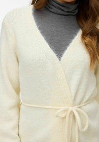 Crèmekleurige overslagvest gemaakt van zacht, pluizig breimateriaal met een ceintuur om de taille en een diep V-hals ontwerp.
