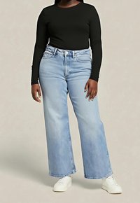 Personne portant un jean large bleu clair, un haut à manches longues noir et des baskets blanches, debout devant un fond uni.