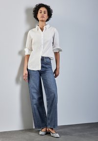 Biała koszula z guzikami z podwiniętymi rękawami, połączona z szerokimi niebieskimi dżinsami i ozdobnymi balerinami. Gładkie tekstury i swobodny styl.