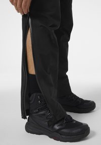 Zwarte waterdichte broek met full-length zijritsen, gecombineerd met zwarte wandelschoenen met veters en robuuste zolen.