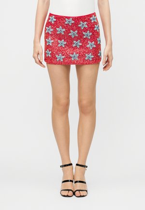 Γυναίκα που φοράει κόκκινη μίνι φούστα διακοσμημένη με μπλε παγιέτες σε σχήμα αστεριού και μαύρα ψηλοτάκουνα πέδιλα με λουράκι στον αστράγαλο.