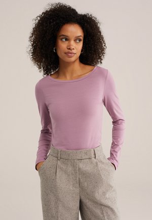 Jeune femme aux cheveux bouclés portant une chemise à manches longues violet clair et un pantalon beige taille haute, les mains dans les poches.