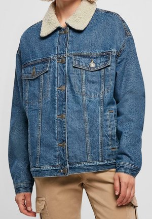 Mężczyzna ubrany w niebieską kurtkę dżinsową z białym kołnierzem z polaru i beżowe spodnie typu cargo, stojący na jednolitym tle.