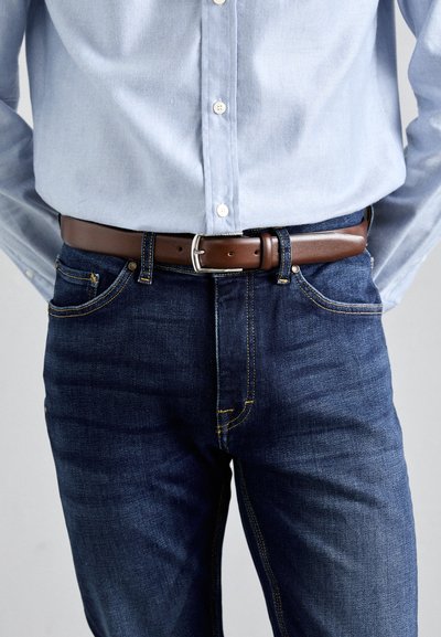 Blue denim jeans with yellow stitching, featuring a brown leather belt and silver buckle, paired with a light blue button-up shirt.
