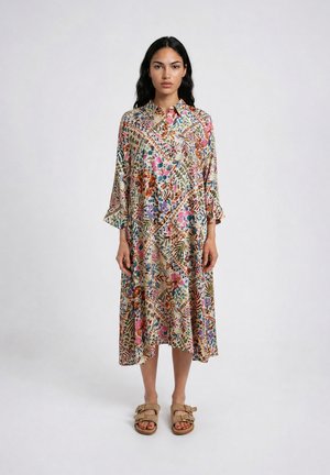 Woman standing straight wearing a colorful floral patterned long-sleeve midi dress and beige sandals with buckles on a plain background.