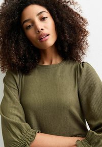 Haut vert olive en tissu texturé, col rond et manches bouffantes, avec des détails froncés au niveau des poignets. Fond neutre.