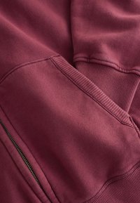 Gros plan sur un tissu de sweat-shirt bordeaux montrant un poignet côtelé, une poche avant avec une couture visible, et une partie d'une fermeture éclair.