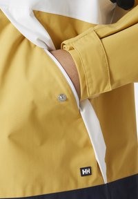 Giacca gialla con un pannello bianco, dotata di un bottone a pressione, tasche laterali e un patch con logo. Il tessuto appare liscio e resistente all'acqua.