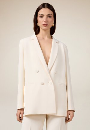 Blazer doppiopetto color crema con scollatura a V, grandi bottoni, silhouette strutturata e design minimalista. Texture del tessuto liscia.