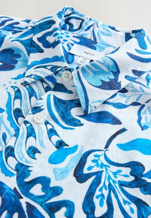 Chemise à manches courtes avec un motif floral bleu sur fond blanc, fabriquée dans un tissu texturé et dotée de deux boutons de fermeture au col.