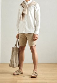 Pantaloni corti beige, camicia bianca a maniche lunghe, leggero pullover gettato sulle spalle, che porta una borsa a tracolla beige, indossando sandali aperti marroni.