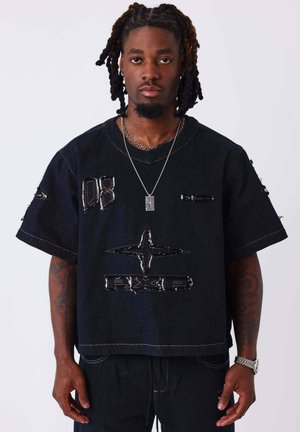 Hombre con rastas que lleva una camiseta negra de gran tamaño con estampados gráficos, collar plateado con cruz, reloj y tatuajes en los brazos sobre un fondo blanco.