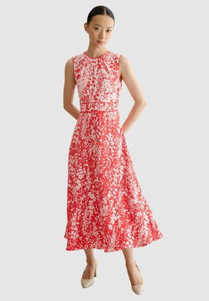 Femme portant une robe midi rouge sans manches à imprimé floral blanc et des chaussures beiges à talons bas, debout les mains derrière le dos sur un fond lumineux.
