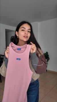 Young woman with long dark hair holds up a pink sleeveless top labeled "HUGO" inside a bright, minimally decorated room.
