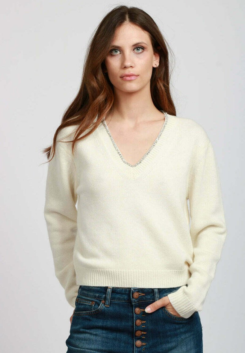Donna con lunghi capelli castano indossa un maglione a V color crema con rifiniture argentate, abbinato a jeans blu scuro a vita alta con bottoni, mani in tasca.