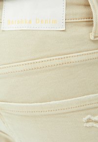 Tela de mezclilla beige con costura doble, un pequeño detalle rasgado y una etiqueta blanca que dice "Bershka Denim" en texto amarillo.
