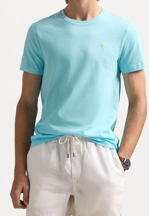 Homme portant un t-shirt à manches courtes bleu clair avec un petit logo jaune sur la poitrine et un pantalon blanc à cordon, avec une montre au poignet gauche.