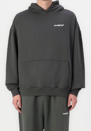 Osoba ubrana w ciemnoszarą bluzę z kapturem i pasujące spodnie z małymi białymi logo „lowlights” na klatce piersiowej i udzie.