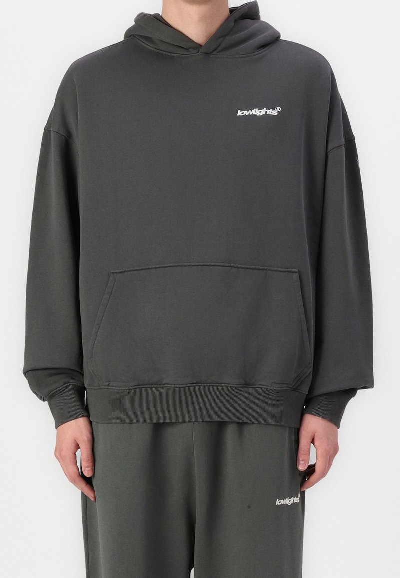 Personne portant un sweat à capuche gris foncé et un pantalon assorti avec de petits logos blancs "lowlights" sur la poitrine et la cuisse.