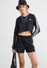 Svart kortermet langermet topp med tre hvite striper på ermene, sammen med svarte shorts med sidelommer og matchende logo detaljer.