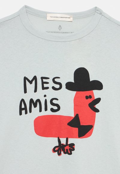 Ανοιχτό μπλε πουκάμισο με ένα κόκκινο καρτούν πουλί που φοράει μαύρο καπέλο και παπιγιόν, με το κείμενο "MES AMIS" πάνω από το πουλί.