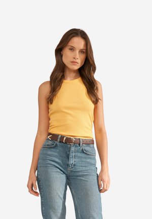 Femme aux longs cheveux bruns portant un haut jaune sans manches, un jean bleu taille haute et une ceinture marron, debout devant un fond blanc uni.