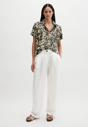 Frau trägt ein kurzärmeliges, beiges Blumenhemd mit weißen, hochtaillierten, weitem Hosenbeinen und beigen Sandalen vor einem einfarbigen Hintergrund.