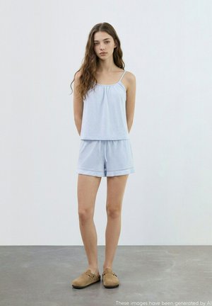 Giovane donna con capelli castani lunghi che indossa una canottiera azzurra chiara e pantaloncini abbinati, in piedi all'interno su pavimento grigio contro una parete bianca.