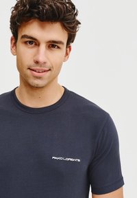 Granatowa bawełniana koszulka z okrągłym dekoltem i krótkimi rękawami. Posiada małe białe logo wyszyte na lewej piersi. Gładka tekstura.