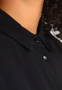 Czarna koszula z kołnierzykiem o gładkiej fakturze, z widocznym zapięciem na guziki oraz małym detalem guzika. Materiał wydaje się lekki i przewiewny.