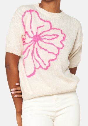 Roomkleurige trui met korte mouwen en een groot roze bloemmotief, gedragen met witte broek, model poseert met hand op de tegenovergestelde arm rustend.