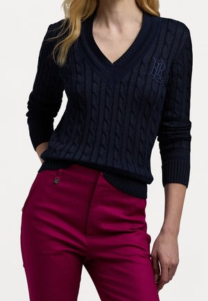 Vrouw die een marineblauwe kabelgebreide V-hals trui draagt en magentakleurige hoge taille broek, staand met één hand in haar zak.
