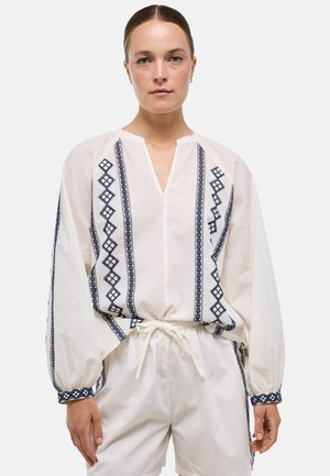 Femme portant un chemisier blanc avec une broderie géométrique bleue et un short blanc à cordon, debout devant un fond uni.