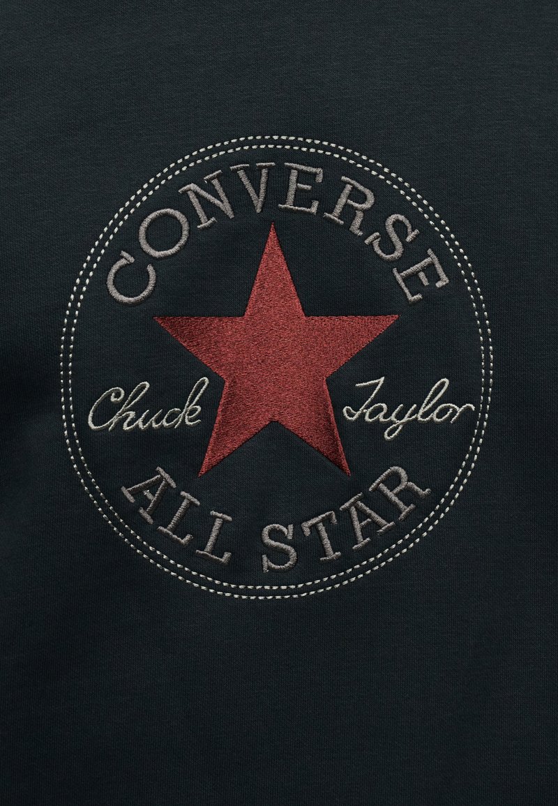 Circular Converse All Star logo with a red star in the center and text "Chuck Taylor" embroidered on dark fabric.
