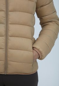 Béžová pufová bunda so zipsom vpredu, horizontálnym prešívaním, elastickými manžetami a bočnými vreckami. Hladká, polstrovaná textúra.