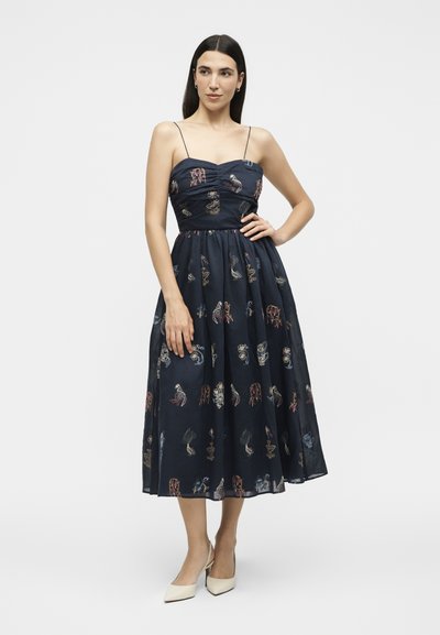 Femme aux cheveux foncés portant une robe midi bleu marine à fines bretelles et motifs abstraits multicolores, associée à des talons pointus blancs.