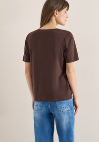 Femme aux cheveux bruns raides portant un T-shirt marron uni à manches courtes et un jean en denim bleu, vue de dos devant un mur beige.