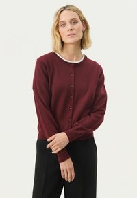 Burgunderfarbener Cardigan aus weichem Strickstoff mit langen Ärmeln, Rundhalsausschnitt und Knopfverschluss vorne. Kombiniert mit einer weißen Unterlage.