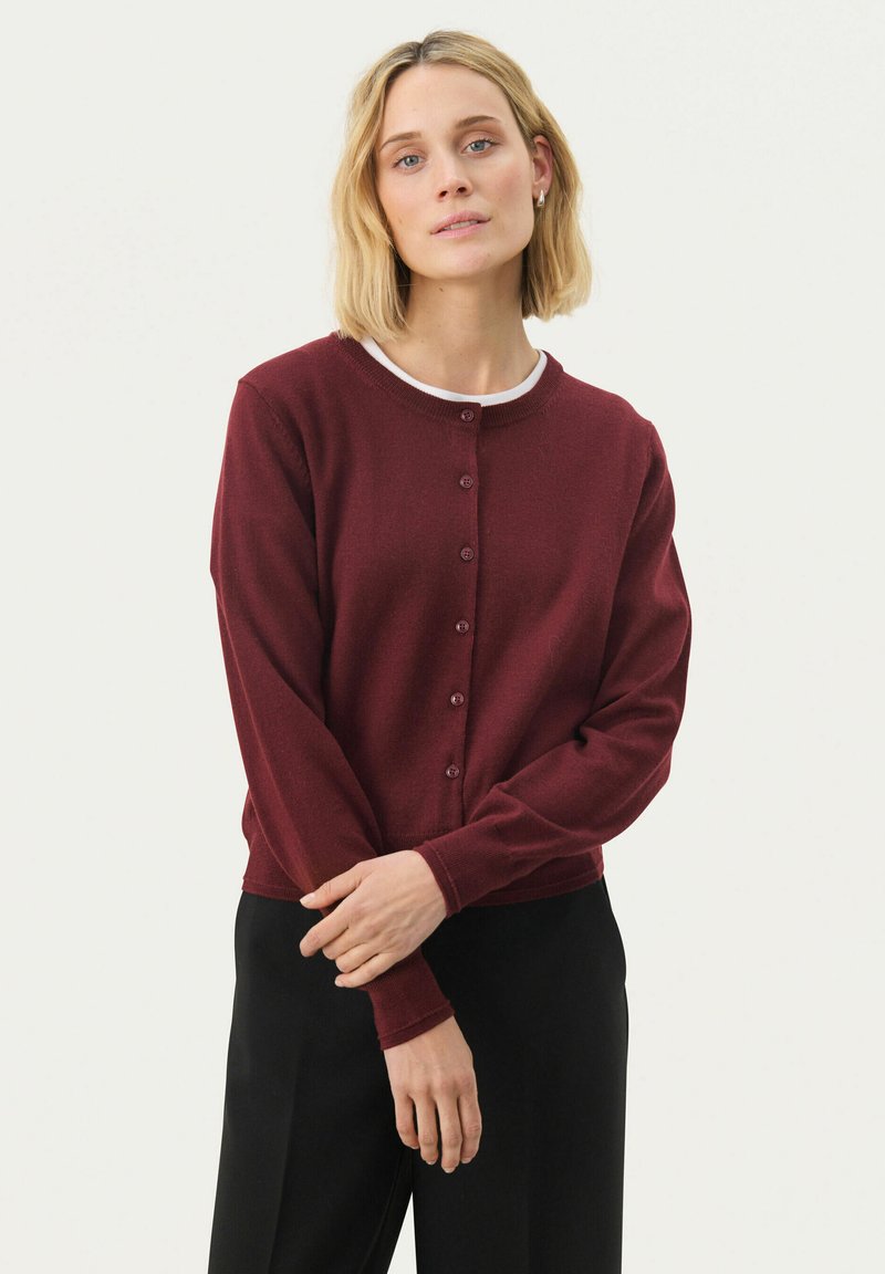 Burgunderfarbener Cardigan aus weichem Strickstoff mit langen Ärmeln, Rundhalsausschnitt und Knopfverschluss vorne. Kombiniert mit einer weißen Unterlage.